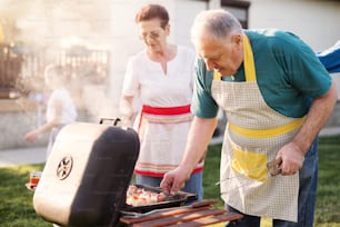 Una pareja de ancianos felices está revisando la carne en una barbacoa mientras su familia disfruta en un patio trasero