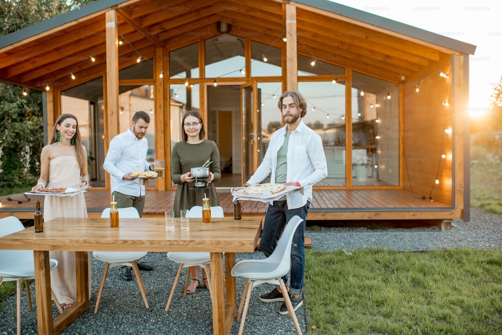 Happy friends carrying food preparing for the dinner on the backyard of the modern wooden house during the sunset