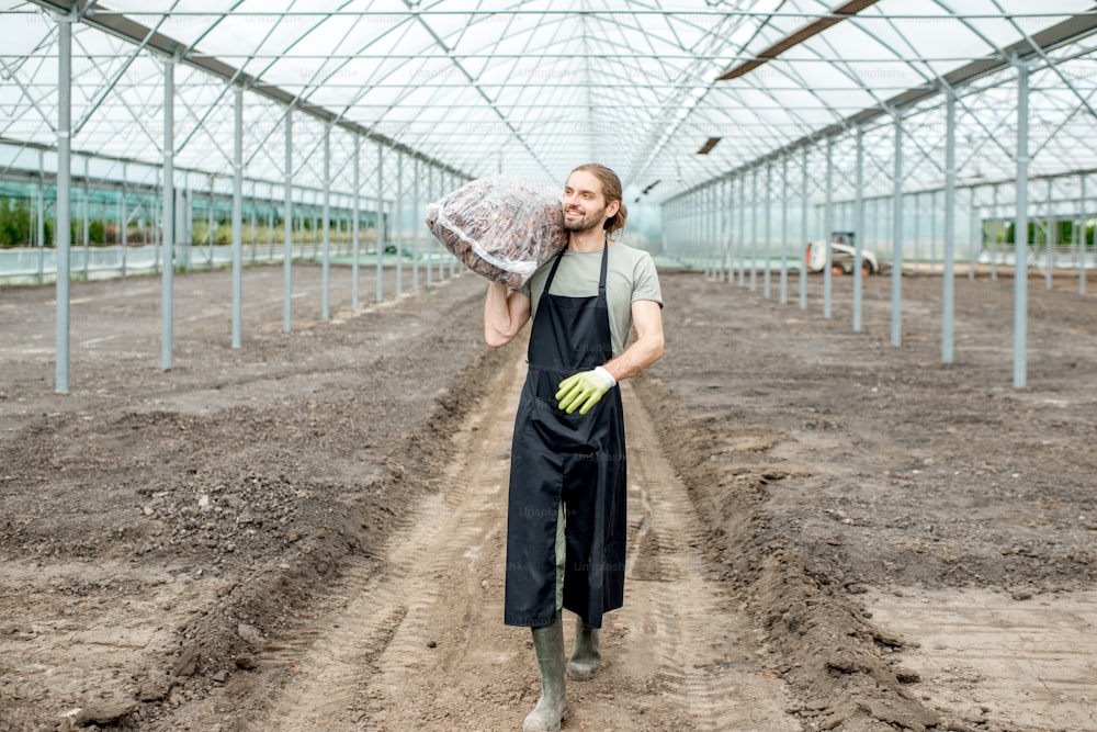 Granjero guapo con uniforme de trabajo que lleva una bolsa con corteza para cubrir con mantillo en el invernadero