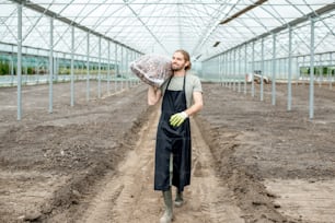 Handsome farmer in working uniform carrying bag with bark for mulching in the glasshouse