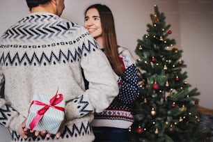 Mann hält Überraschungsgeschenk hinter dem Rücken für seine Frau. Weihnachten tauscht Geschenke aus. Familie glückliche Momente. Frohe Weihnachten und ein glückliches neues Jahr, saisonale Grüße. Frohe Feiertage