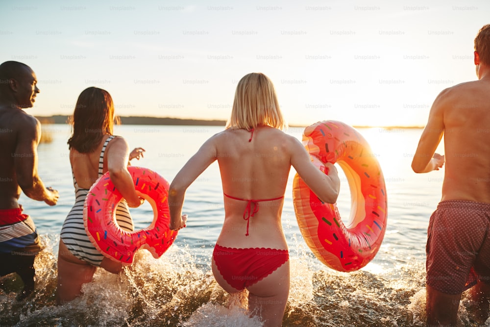 Vista traseira de um grupo de diversos amigos em trajes de banho espirrando água enquanto correm para um lago juntos em uma tarde de final de verão