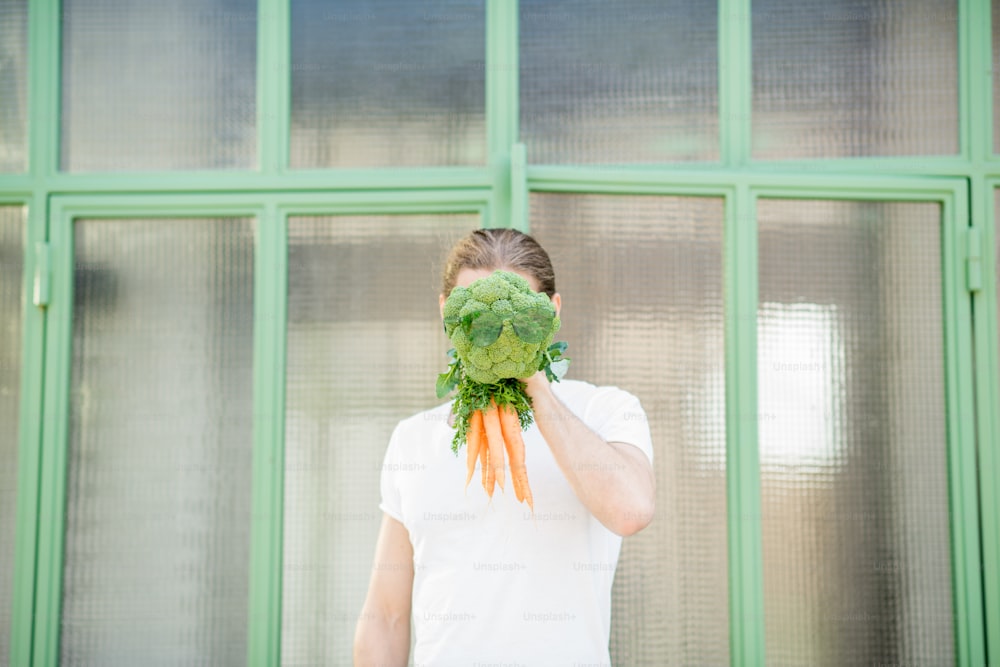 Retrato engraçado de um homem segurando brócolis com cenoura em vez de sua cabeça ao ar livre. Conceito de alimentação saudável