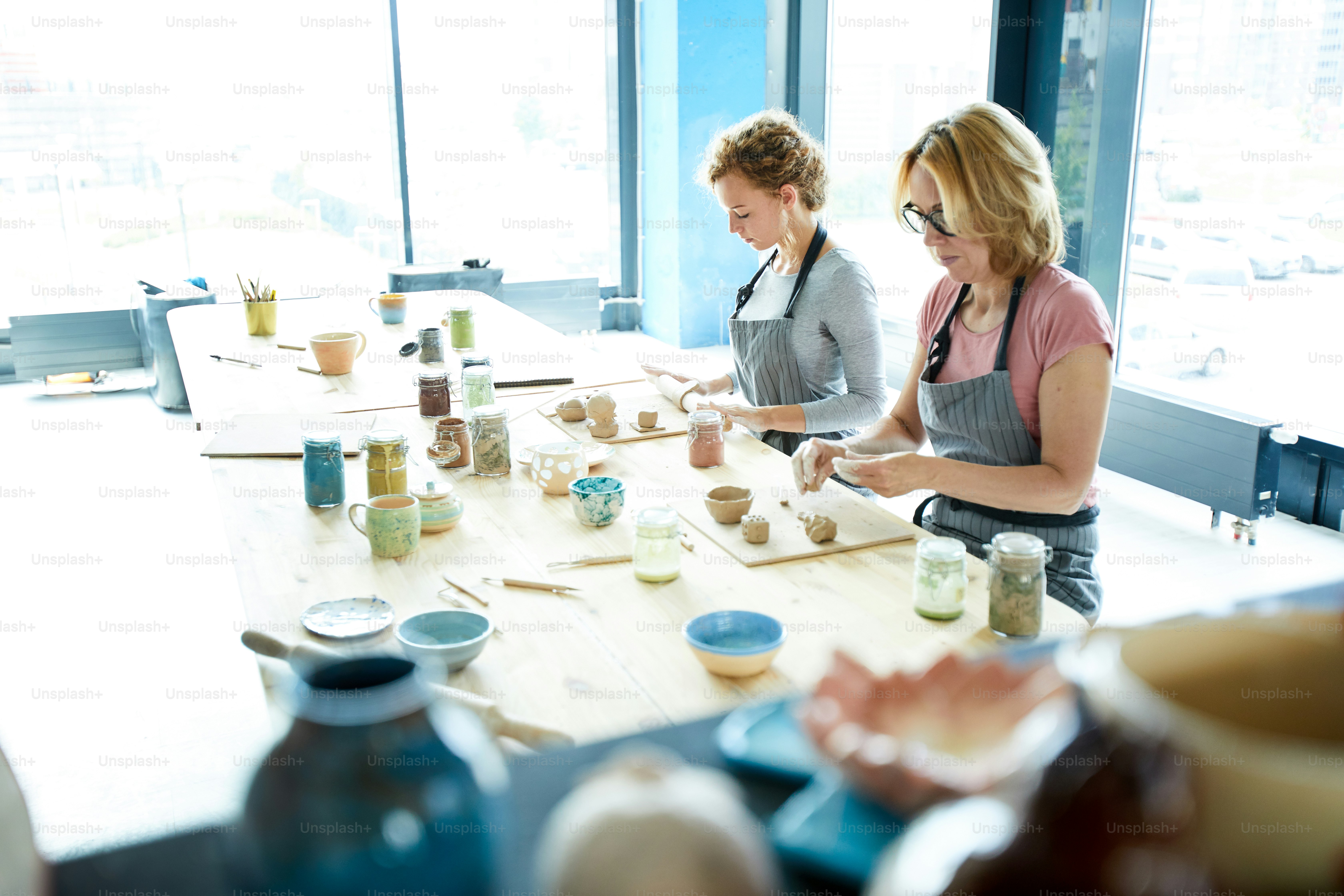 Two women in workwear sitting by table, rolling clay workpieces and creating new items in workshop