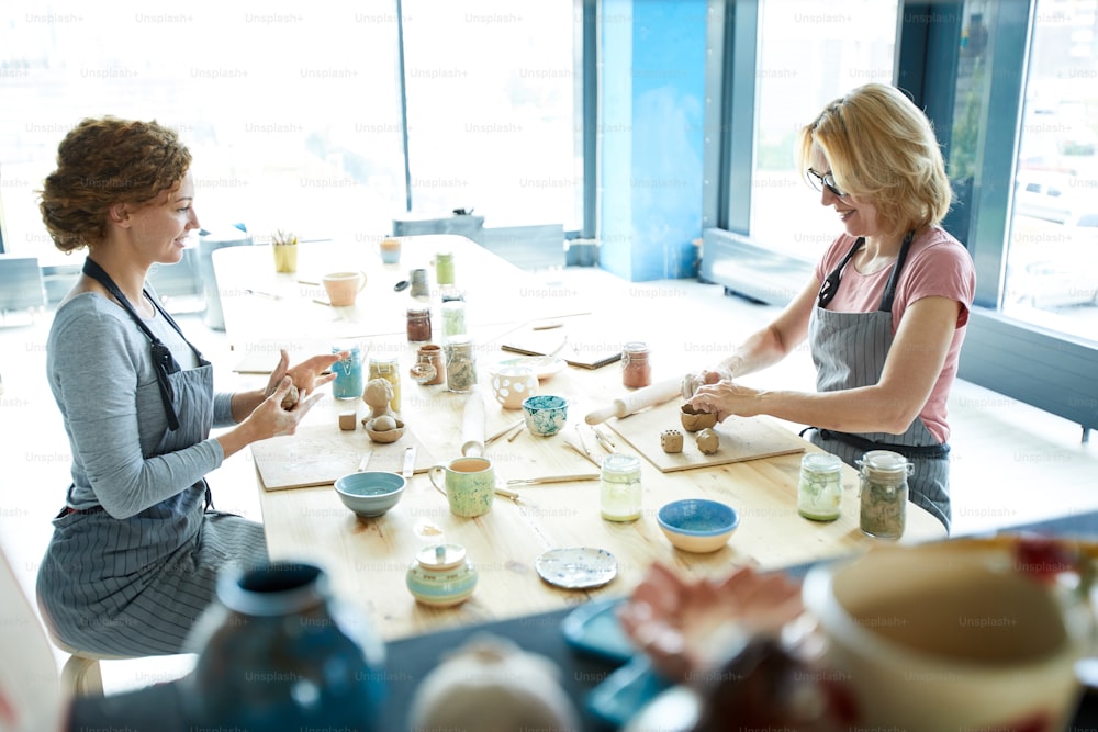Deux femmes créatives en tabliers assises dans un studio d'artisanat en argile et créant leur propre faïence
