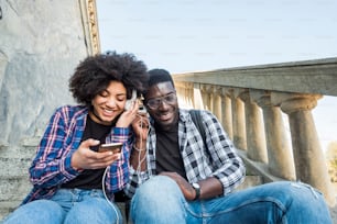couple enjoying the music and the modern technology with phone together in friendship. black race african with afro ethnic hair. smiles and having fun in relationship. young people enjoying at the city