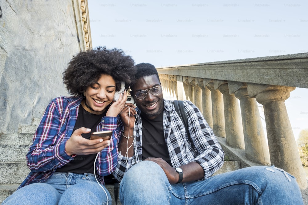 우정으로 함께 전화로 음악과 현대 기술을 즐기는 커플. 아프리카 민족 머리를 가진 흑인 인종. 미소 짓고 관계를 즐깁니다. 도시에서 즐기는 젊은이들