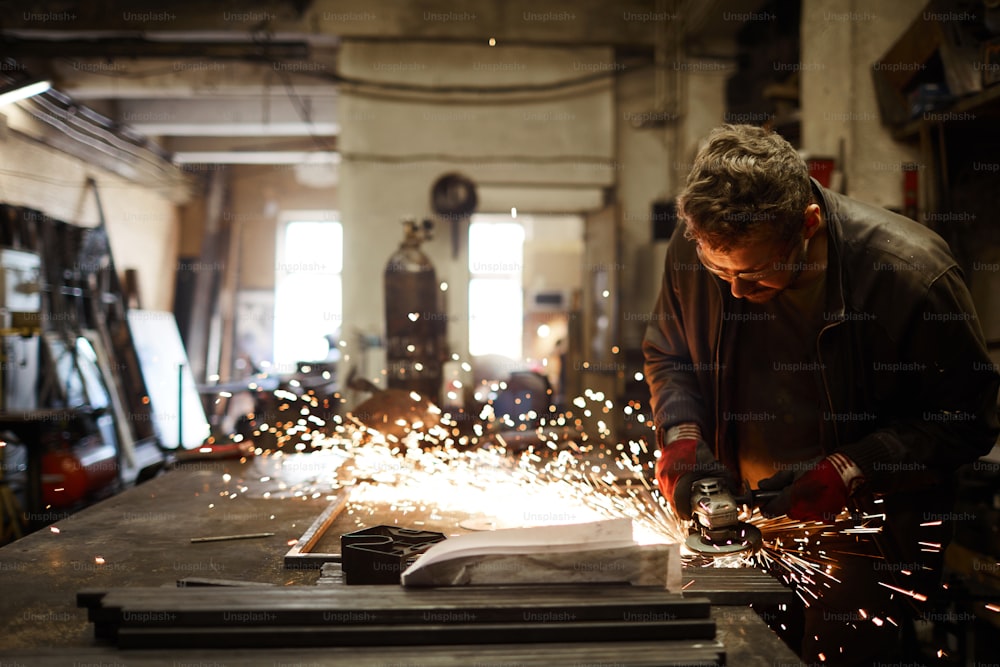 Giovane fabbro contemporaneo in piedi vicino all'incudine di ferro e alla lavorazione del pezzo con l'utensile elettrico