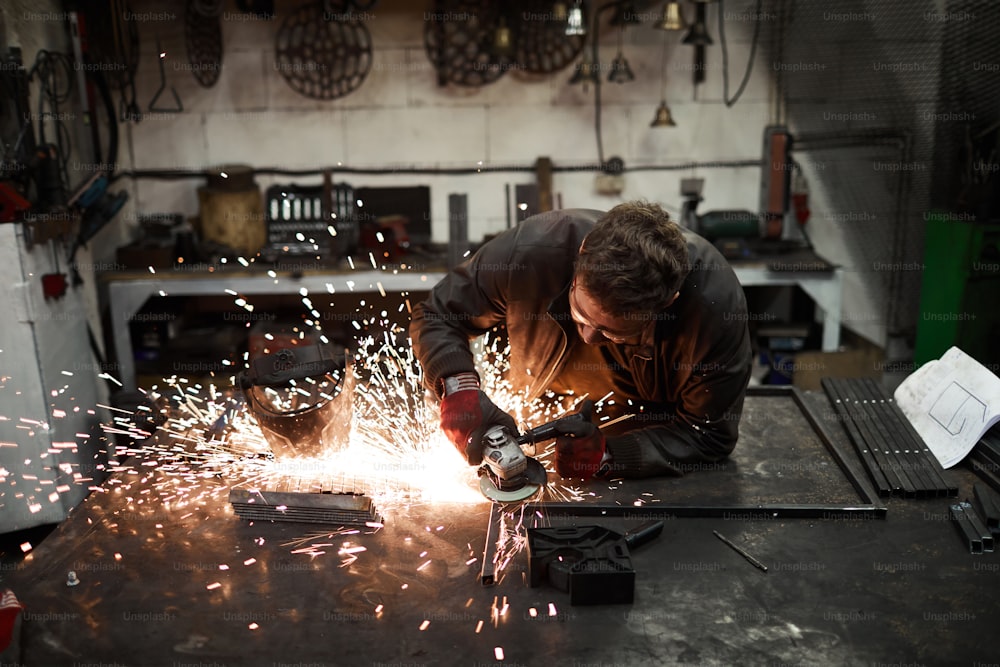 Young blacksmith standing by anvil and welding or processing iron workpiece with electric handtool