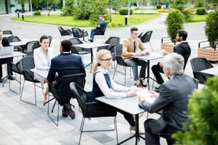 Kleine Gruppen von Mitarbeitern in formeller Kleidung sitzen an Tischen im Sommercafé im Freien, entspannen sich und unterhalten sich