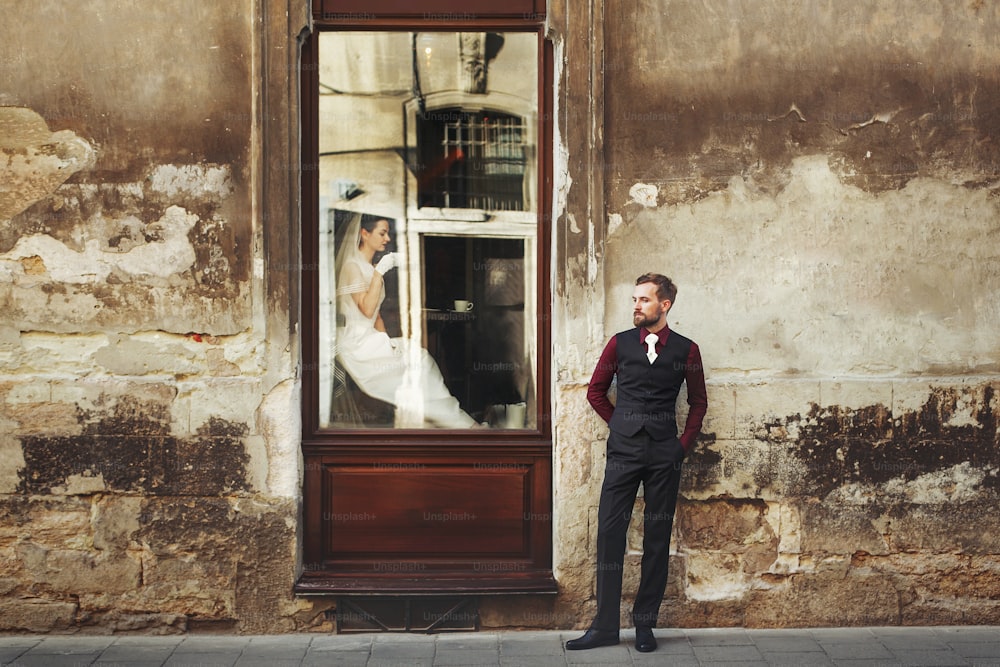 elegant gorgeous bride  drinking coffee in window and stylish groom posing at building in street outdoors cafe. unusual luxury wedding couple in retro style. romantic moment