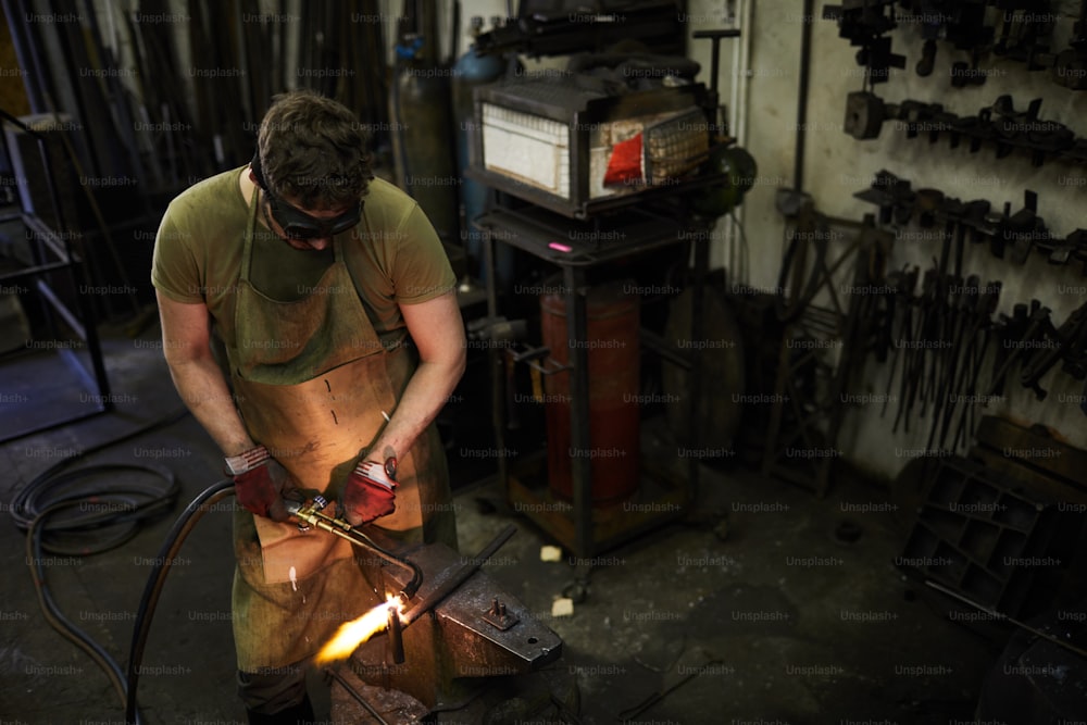 Concentrated busy blacksmith in dark safety goggles moving torch while cutting metal on anvil, he welding in workshop