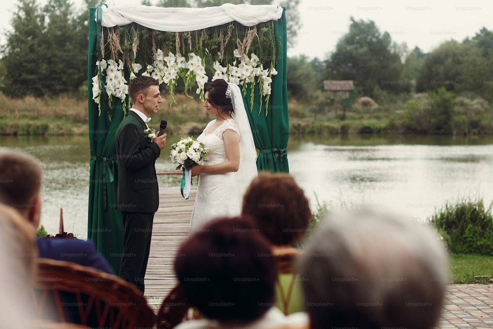 スタイリッシュなスーツを着たハンサムな感情的な新郎は、通路と湖の近くの屋外の結婚式で花束を持つ美しい花嫁に結婚式の誓いを立て、前景のゲスト