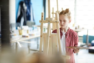 Serious youthful girl with color palette looking at painting on her easel while standing in front of it
