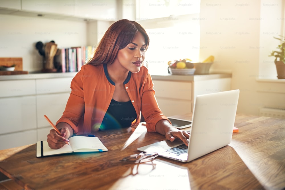 Young African female entrepreneur sitting at a table in her kitchen writing down notes and working on a laptop