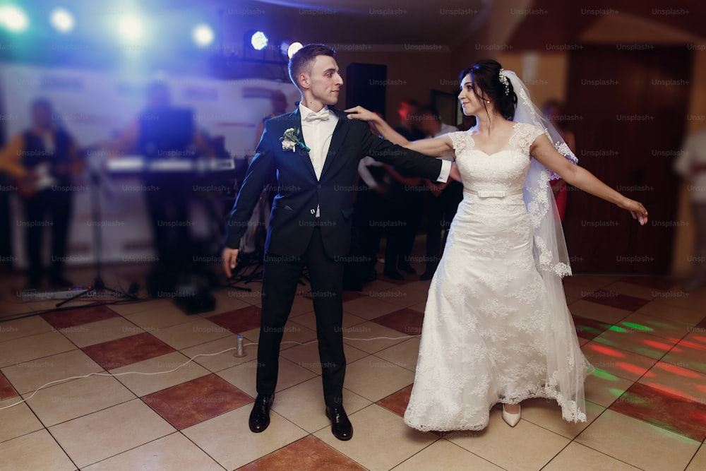 Romantic newlywed couple dancing, handsome groom and beautiful happy bride first dance while holding hands at restaurant wedding reception, beautiful lighting