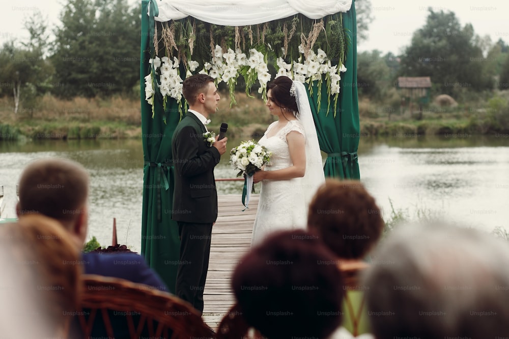 통로와 호수 근처의 야외 결혼식에서 꽃다발로 아름다운 신부에게 결혼 서약을 하는 세련된 정장을 입은 잘생긴 감성 신랑, 전경에 손님