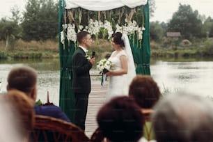 Handsome emotional groom in stylish suit giving wedding vow to beautiful bride with bouquet at outdoor wedding ceremony near aisle and lake, guests in the foreground
