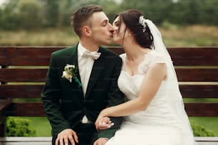 Cute newlywed couple sitting on a bench in the park, smiling handsome groom in stylish suit kissing beautiful happy bride in white dress while holding hands, family portrait