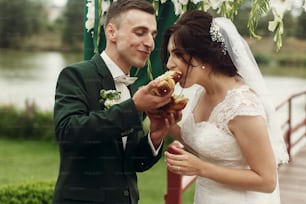 結婚式でパンを割る幸せな新婚カップル、スラブの伝統の屋外で結婚式の通路の近くでパンを食べるハンサムな花婿と笑顔の花嫁