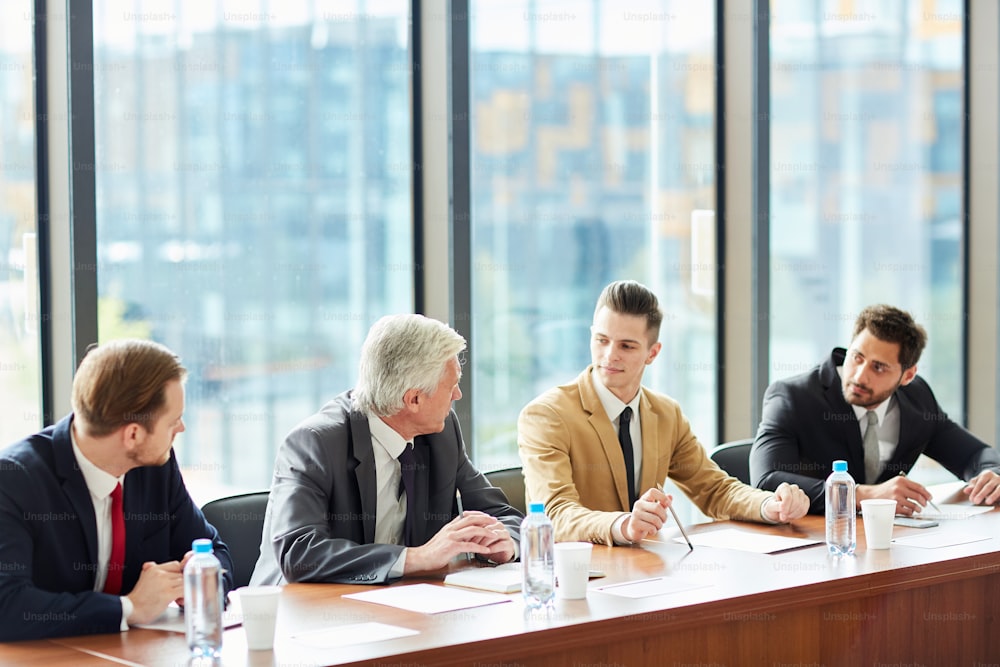 Modern confident business executives in formal suits sitting at conference table and arguing at meeting while discussing company development