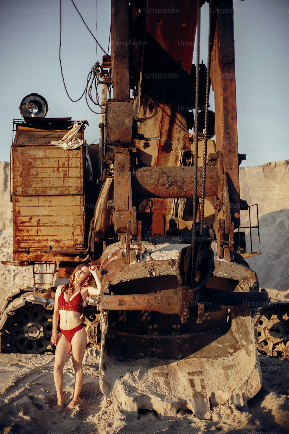 beautiful girl in red swimsuit walking on sandy beach near old machinery in sunset light. attractive young woman in bohemian look with windy hair, posing at bulldozer