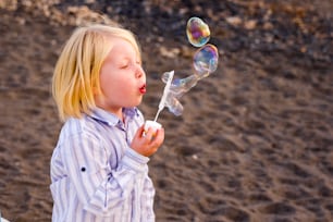 Los niños caucásicos bonitos y hermosos juegan con el jabón de burbujas en la actividad de ocio al aire libre. labios rojos y cabello rubio. playa en el fondo tiempo de vacaciones. Soplar el jabón de burbujas