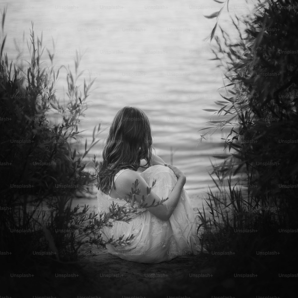 Muchacha triste sentada en la playa de arena a la luz del atardecer cerca del lago. Espalda de mujer joven con vestido bohemio blanco con cabello ventoso cerca del agua. Concepto de soledad y depresión.