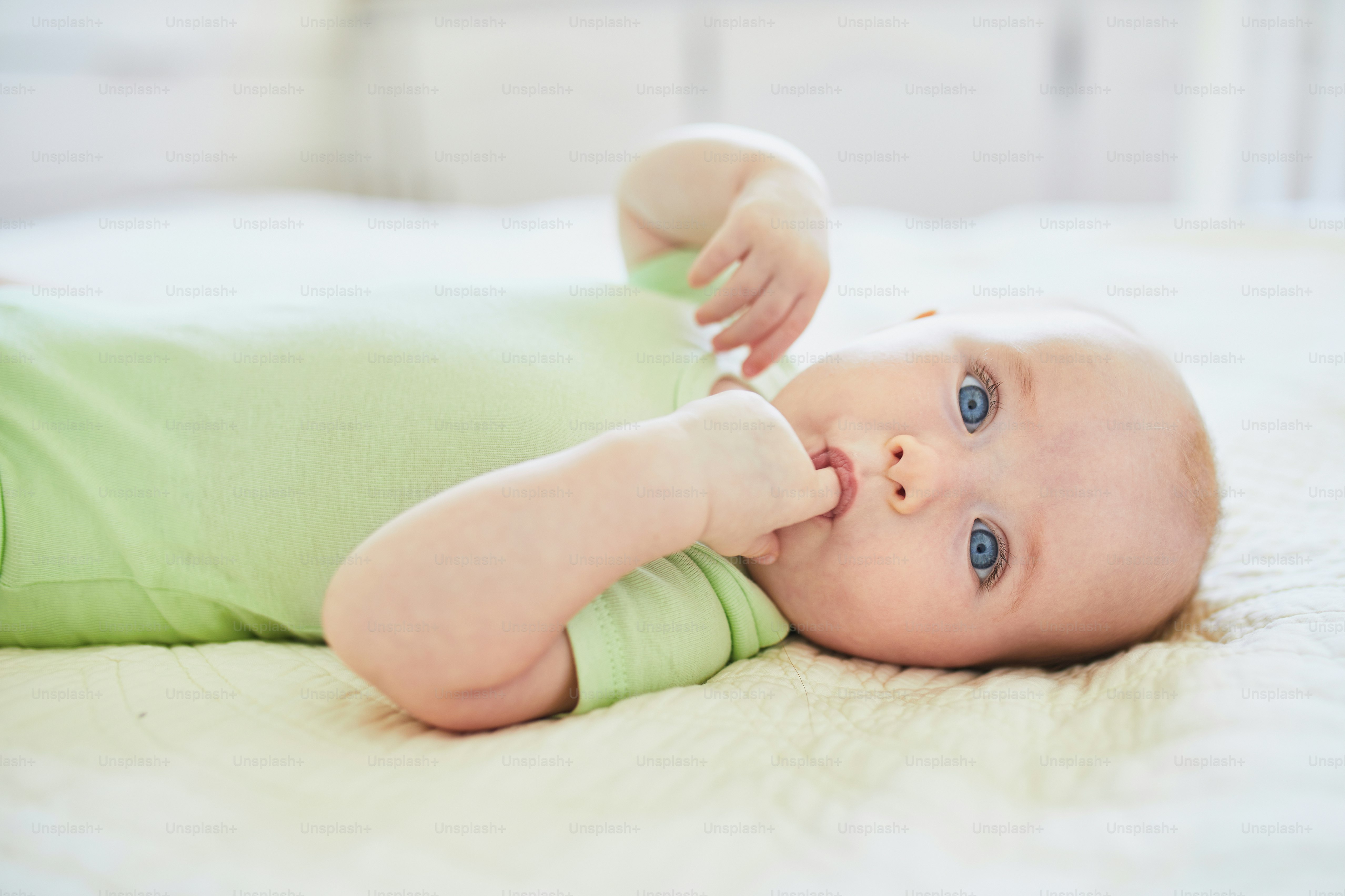 Baby girl suckling her finger to comfort herself. Little child lying on bed. Infant kid in sunny nursery