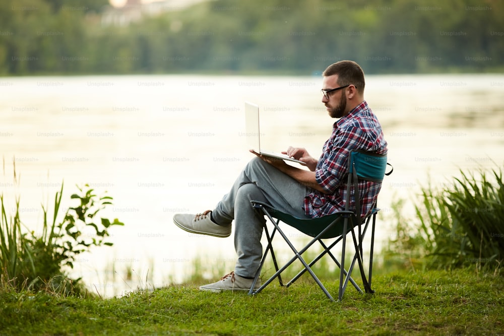 Young man wearing eyeglasses sitting on chair and working on his laptop near the lake on fresh air