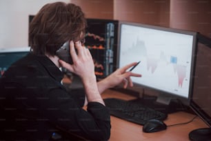 Over the shoulder view of and stock broker trading online while accepting orders by phone. Multiple computer screens ful of charts and data analyses in background.