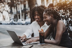 Interracial couple: black laughing girl and Asian smiling guy are looking through funny photos using the laptop while sitting in a street cafe, the glass of ice tea and the smartphone near the girl