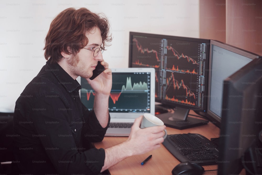 Over the shoulder view of and stock broker trading online while accepting orders by phone. Multiple computer screens ful of charts and data analyses in background.