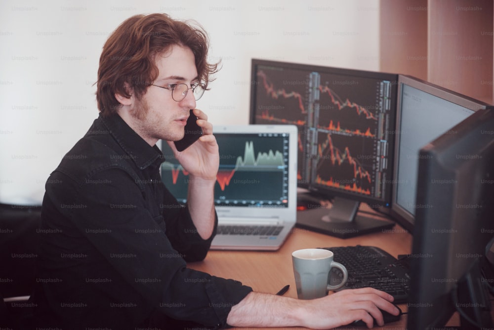 電話で注文を受け付けながらオンラインで取引する株式ブローカーの肩越しの眺め。バックグラウンドでチャートとデータ分析でいっぱいの複数のコンピューター画面。