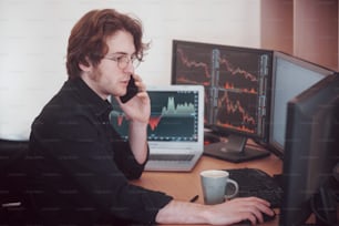 Over the shoulder view of and stock broker trading online while accepting orders by phone. Multiple computer screens ful of charts and data analyses in background.