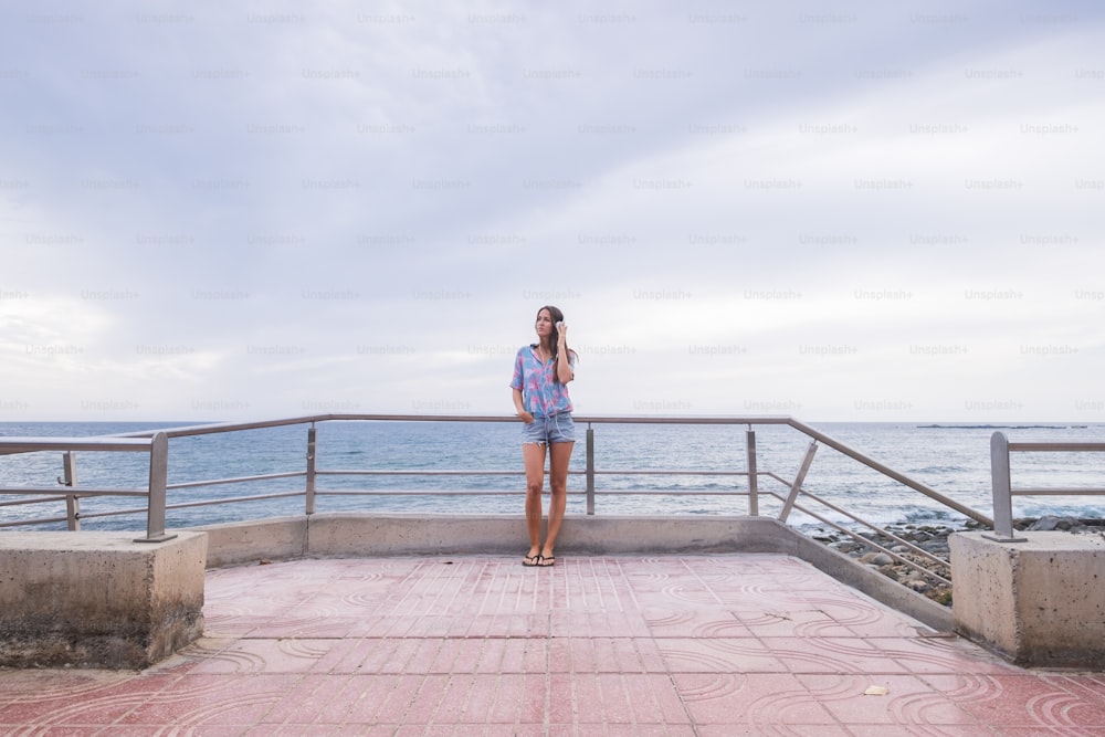 Retrato de cuerpo entero de una hermosa mujer joven de pie con el océano y el agua azul en el fondo y el cielo escuchando música con auriculares. Concepto moderno y actividad de ocio para una chica agradable en la playa