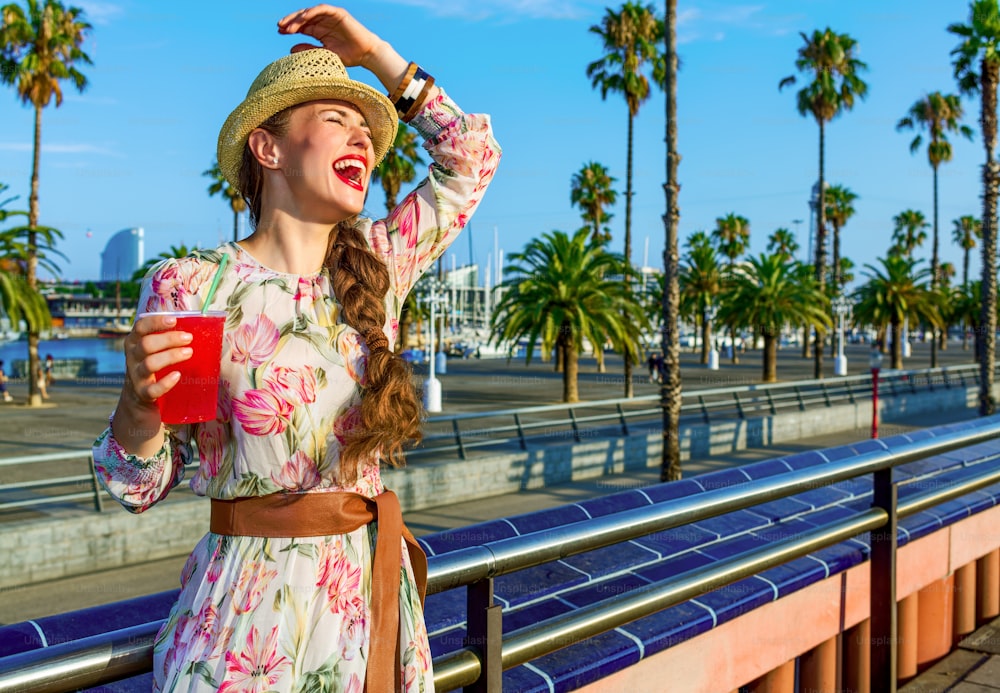 Summertime at colorful Barcelona. happy young woman in long dress and straw hat in Barcelona, Spain with bright red beverage having fun time