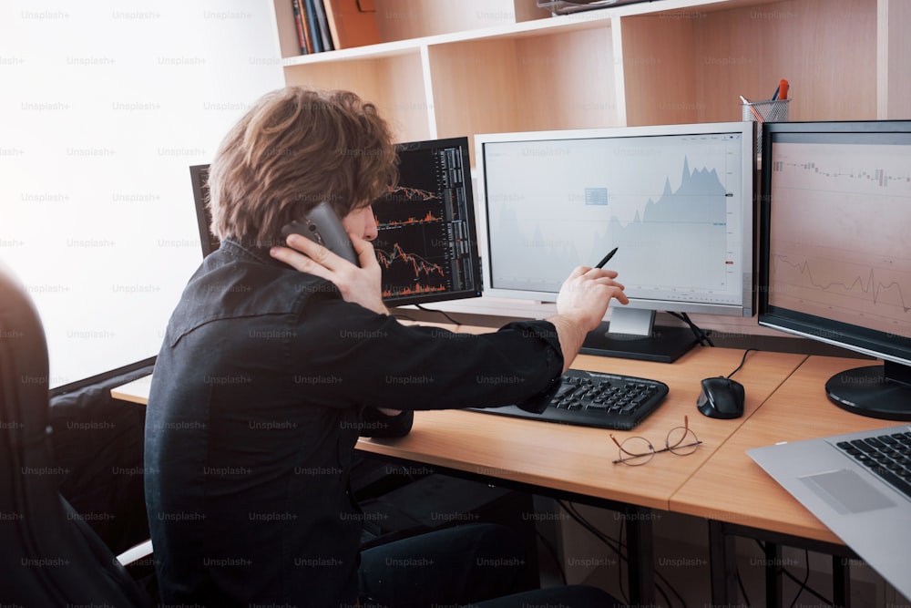 Over the shoulder view of and stock broker trading online while accepting orders by phone. Multiple computer screens ful of charts and data analyses in background.