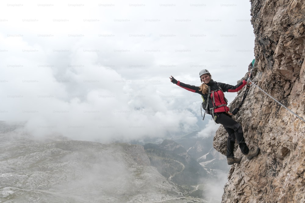 young attractive female university student on a vertical and exposed rock face climbs a Via Ferrata while smiling and waving and making a peace sign