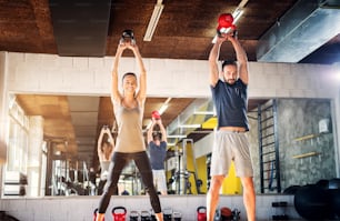 Strong young happy couple lifting kettle bells in a gym. Standing in front of a camera..
