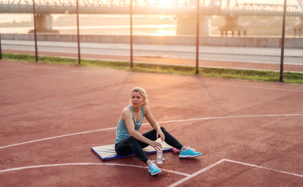 Cansada joven deportiva linda rubia sentada afuera en una colchoneta de entrenamiento después de un duro entrenamiento. Entrenamiento matutino.