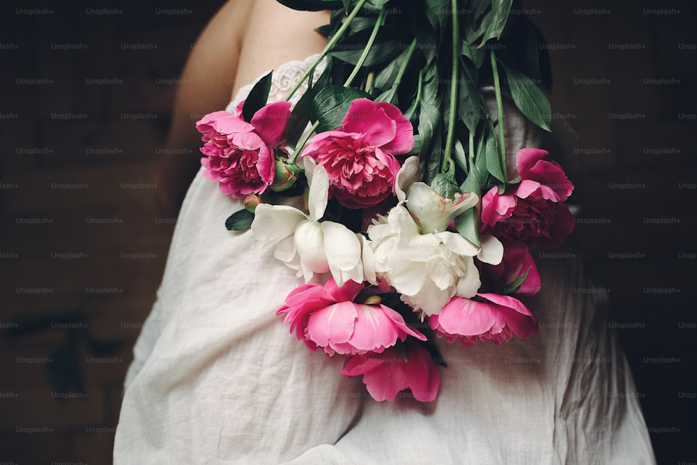 beautiful pink peonies on legs of boho girl in white bohemian dress, top view. space for text. stylish hipster woman sitting with flowers in moody morning. atmospheric sensual moment