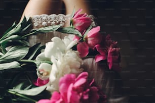 hermosas peonías rosadas en las piernas de la chica boho en vestido bohemio blanco, vista superior. espacio para el texto. Mujer hipster con estilo sentada con flores en la mañana malhumorada. Momento sensual atmosférico