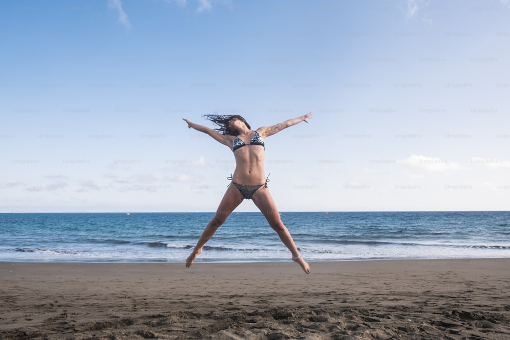 beautiful body fitness lifestyle young woman jump full of happiness at the beach near the shore and the waves of the blue ocean. joyful and summer fun concept for caucasian cheerful people