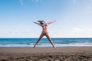 beautiful body fitness lifestyle young woman jump full of happiness at the beach near the shore and the waves of the blue ocean. joyful and summer fun concept for caucasian cheerful people
