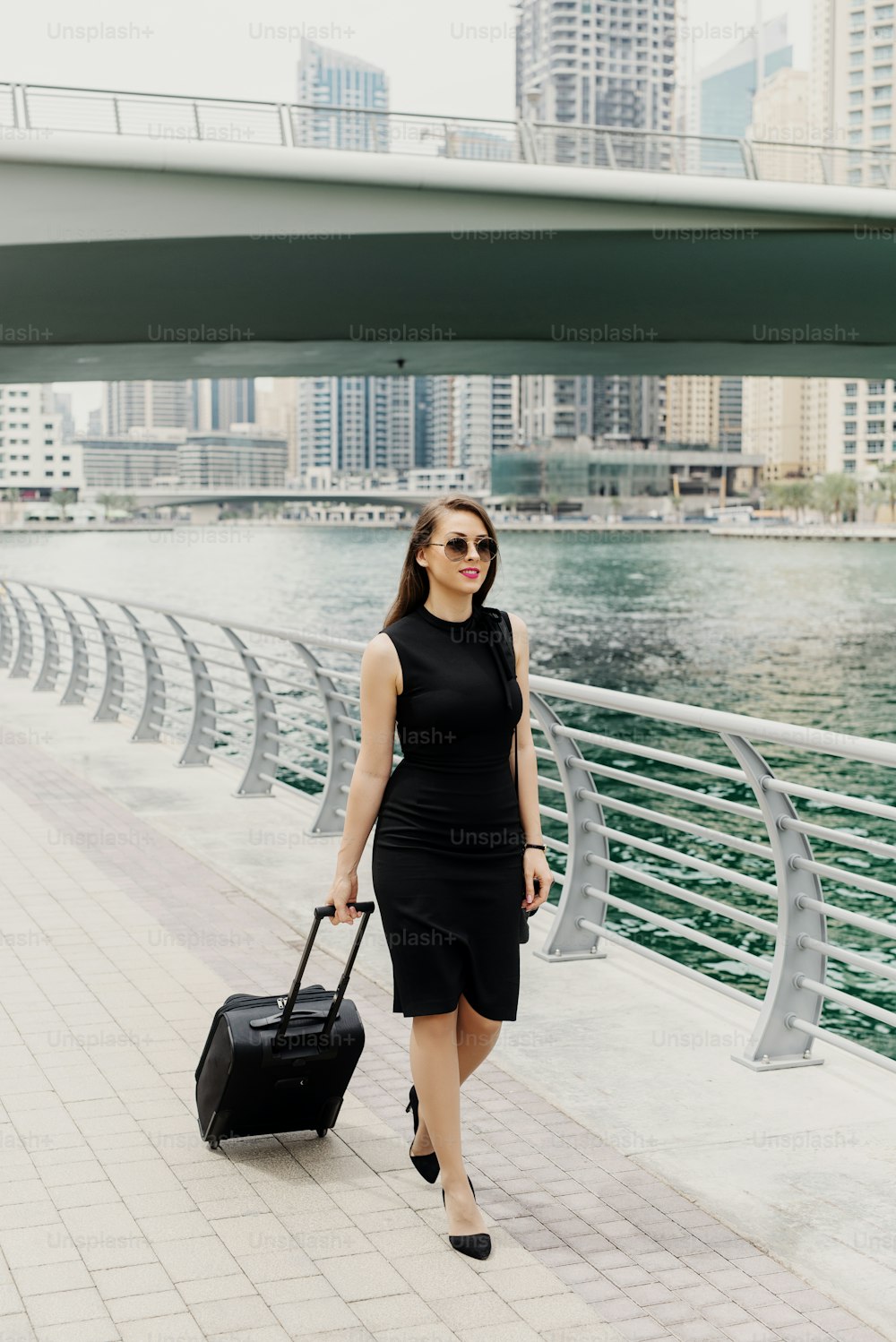Joven mujer de negocios moderna y segura de sí misma tirando de una maleta en un Dubai Marine. Empezar un nuevo trabajo en una gran ciudad.