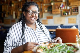 Mujer joven feliz con trenzas africanas mirándote mientras estás sentada junto a la mesa y almorzando pizza