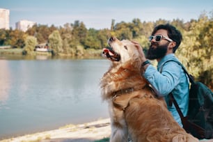 Best friends' concept - between human and animal. Angle view, young man hugging, tapping and sitting with his dog on the chair in the park, enjoying the view of the lake