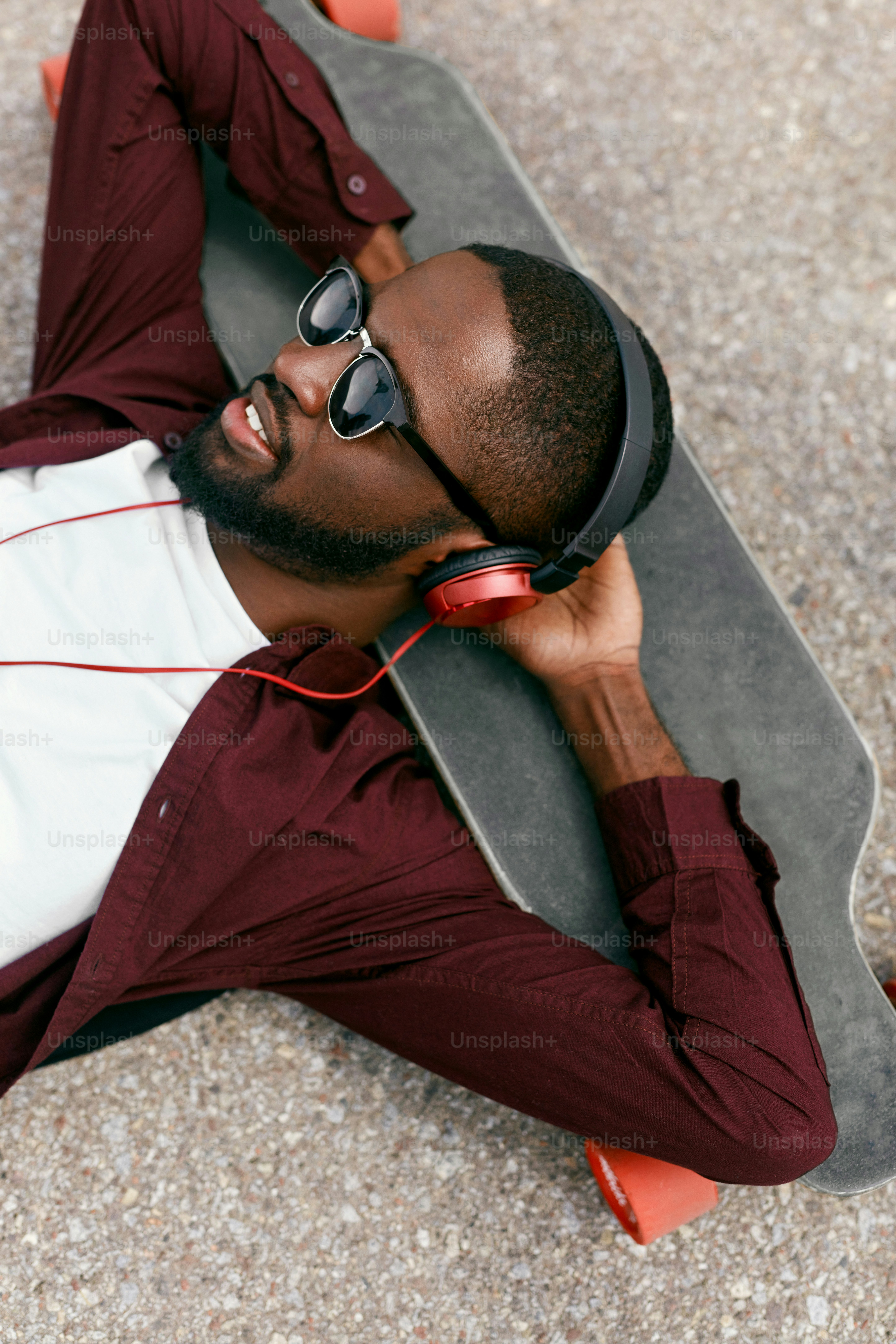 Moda. Homem negro ouvindo música em fones de ouvido na rua, deitado em longboard. Alta Resolução