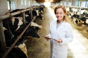 Portrait of cheerful female veterinarian smiling looking at camera while using digital tablet standing in cowshed of modern dairy farm, copy space